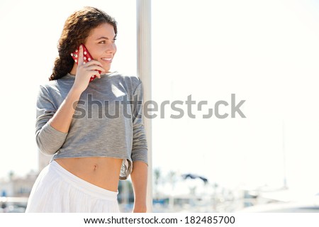 Close up portrait of a beautiful sophisticated young woman on holiday smiling against a sunny sky and using a smartphone to make a call. Travel and technology lifestyle.