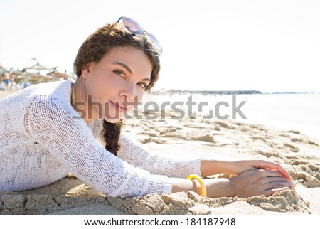 Portrait of an attractive young woman laying down on a golden sand beach on holiday smiling at the camera during a sunny day in a coastal destination. Travel and healthy lifestyle, outdoors.