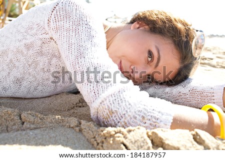 Side portrait of an attractive young woman laying down on a golden sand beach on holiday smiling at the camera during a sunny day in a coastal destination. Travel and healthy lifestyle, outdoors.