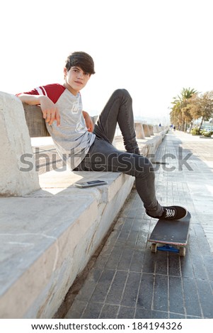 Side view of an attractive and active teenager student relaxing during a sunny day sitting on a bench by the sea with his skateboard and his smartphone. Outdoors technology and lifestyle.