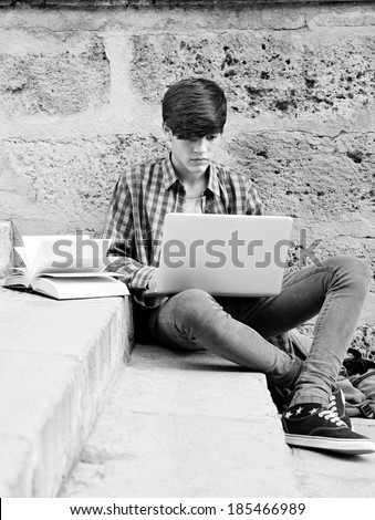 Attractive teenager boy sitting on a college campus with school books and a laptop computer against an old stone building wall doing his homework. Education technology lifestyle.