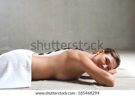 Side view of an attractive and elegant mature woman relaxing laying down with a towel in a health spa having a volcanic black stones heat treatment on her back, smiling. Beauty lifestyle.