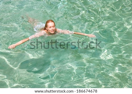 Attractive young woman swimming in the clear and transparent blue waters of a health spa swimming pool, relaxing on a summer holiday trip and enjoying a swim during a sunny day, outdoors.