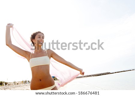 Beautiful young woman visiting a beach on holiday and holding a flying pink silk fabric sarong up against the sky with her arms, joyfully smiling while enjoying a summer day. Beauty and lifestyle.