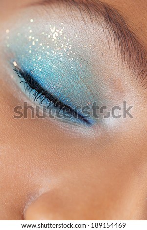 Close up beauty portrait detail of an african american black young woman closed eye wearing bright colorful blue eye make up and glitter for a fun party. Cosmetics, personal care and beauty lifestyle.