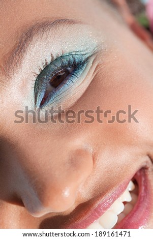 Over head beauty close up portrait of an attractive joyful young black woman half face looking at camera and wearing colorful fantasy make up cosmetics, smiling outdoors. Aspirational lifestyle.
