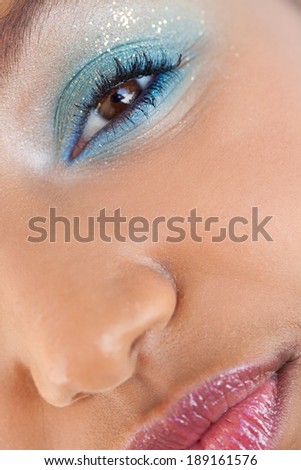 Over head beauty close up portrait of an attractive young black woman half face and eye wearing colorful fantasy make up cosmetics and smiling at the camera, indoors. Aspirational skin perfection.