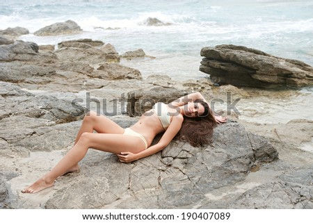 Side body view of a beautiful young woman relaxing and laying on the dark textured rocks of a beach with mountains, lounging and enjoying sunbathing on a vacation by the sea. Beauty and lifestyle.