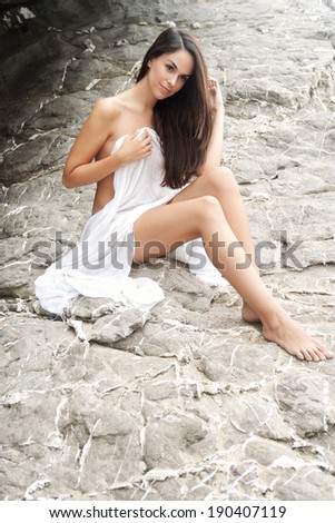 Side view of an attractive young woman relaxing on a gray textured rock face on a beach, wearing a white sarong and being thoughtful and serene during a summer holiday. Beauty and lifestyle outdoors.