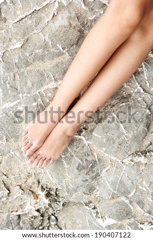 Over head close up beauty detail of a young attractive woman bare legs and feet sitting and relaxing on a textured rock by the sea sunbathing during a summer holiday. Health and beauty lifestyle.