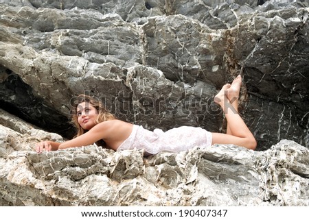 Side view of an attractive young woman laying and relaxing on a gray textured rock face on a beach, in a white sarong and smiling thoughtfully during a summer holiday. Beauty and lifestyle outdoors.