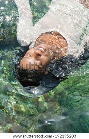 Over head portrait view of an attractive african american woman swimming and floating in a clear blue sea with sun rays reflecting in the water, relaxing on holiday. Healthy travel lifestyle outdoors.