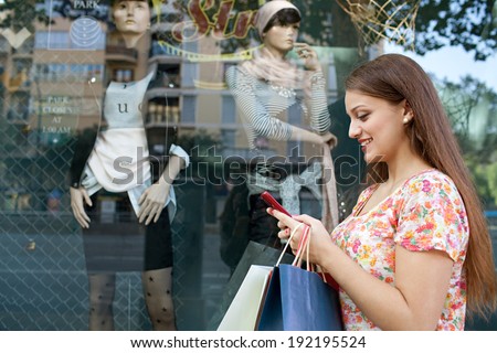 Profile portrait of an attractive joyful woman shopping in a city by a fashion store window with reflections, using a smartphone device to network, outdoors. Consumerism and technology lifestyle.