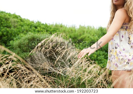 Rear view of a young woman half body relaxing and enjoying a summer holiday, walking in the vegetated dunes of a beach feeling the yellow long grass with her hand. Outdoors healthy travel lifestyle.
