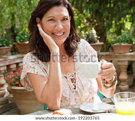 Attractive senior woman at a table having breakfast in a luxury hotel garden during a sunny day on holiday. Mature people eating and drinking healthy food and relaxing, smiling. Outdoors lifestyle.