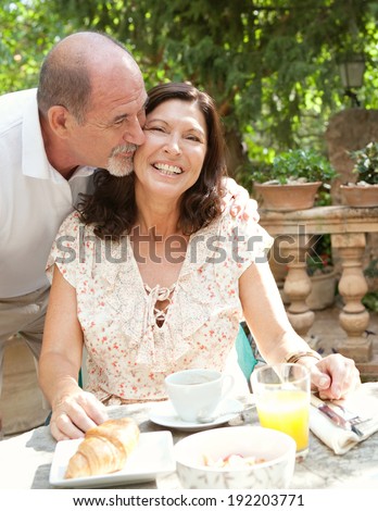 Senior couple having breakfast together at a table in a luxury hotel garden during a holiday. Mature people eating and drinking healthy food enjoying each others company, kissing. Outdoors lifestyle.
