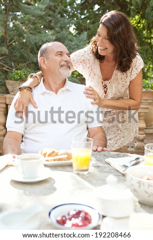 Attractive mature tourists couple having a luxurious continental breakfast in a hotel garden on a sunny day on holiday. Senior people eating healthy food and enjoying retirement, hugging. Lifestyle.