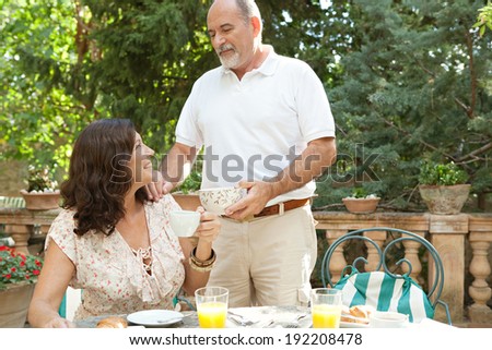 Attractive mature tourists couple having a luxurious continental breakfast in a hotel garden on a sunny day on holiday. Senior people eating healthy food and enjoying retirement, smiling. Lifestyle.