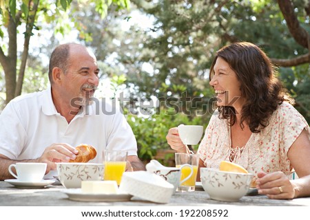 Senior couple having breakfast together at a table in a luxury hotel garden during a sunny day. Mature joyful people eating healthy food and having fun while drinking coffee. Outdoors lifestyle.