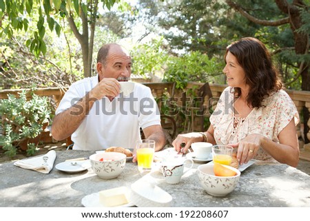 Senior couple having breakfast together sitting at a table in a luxury hotel garden on holiday. Mature joyful people eating healthy food, having a conversation drinking coffee. Outdoors lifestyle.