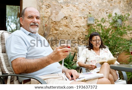 Mature couple sitting at a table in a luxury hotel garden on holiday drinking wine and reading relaxing together on a summer vacation. Senior people enjoying travel and retirement, outdoors lifestyle.