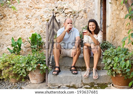 Portrait of a senior man and woman sitting on the stone steps of luxury hotel garden during a sunny day on holiday drinking wine together and relaxing on vacation. Mature people, outdoors lifestyle.
