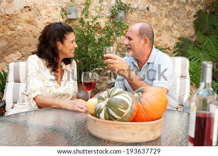 Side portrait of a senior couple relaxing in the garden of a luxury hotel on holiday, enjoying a glass of wine, having a conversation. Mature people drinking at a table, outdoors lifestyle.