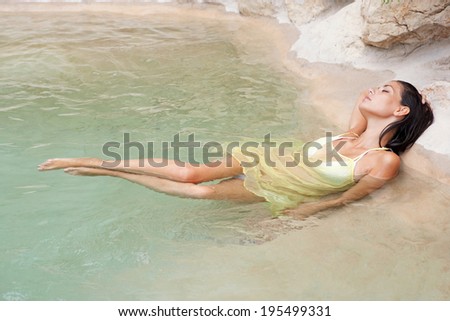 Space view of an attractive young woman laying down on the edge of a spa natural swimming pool submerged in the water and relaxing while on a summer holiday trip. Beauty and healthy lifestyle.