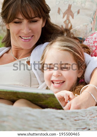 Close up portrait of a beautiful young mother and her five years old daughter laying down in a hammock playfully reading a children stories book during a sunny summer day. Family lifestyle outdoors.