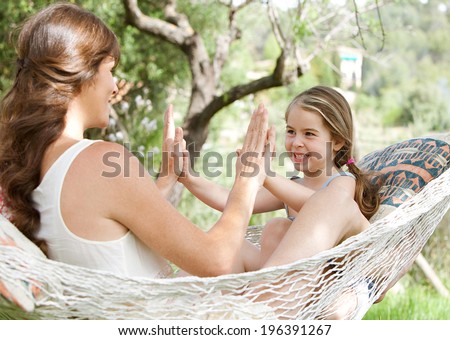 Young daughter and mother sitting together on a hammock in a holiday home garden playing games and clapping their hands, having fun during a sunny summer day on vacation. Active family lifestyle.
