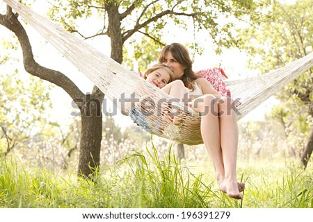 Wide view of a young mother and daughter relaxing together and smiling sitting in a hammock, hugging and lounging during a sunny summer day in a holiday home garden with grass and trees, lifestyle.