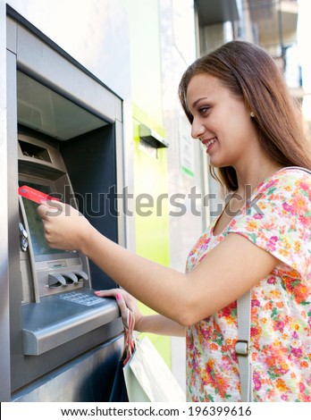 Side portrait view of an attractive young woman using a cash machine to withdraw cash money with her credit card, carrying shopping bags, smiling in a city street. Outdoors finance and lifestyle.
