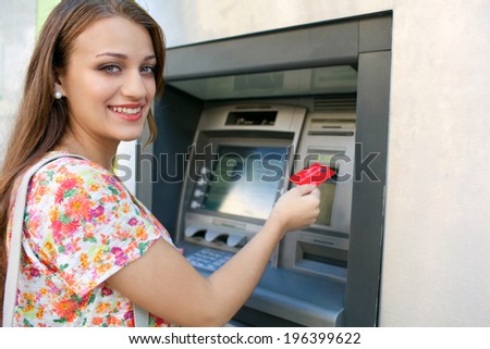 Side portrait view of an attractive young woman using a cash machine to withdraw cash money with her credit card, turning and smiling in a city street. Outdoors finance and lifestyle.