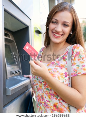 Joyful portrait view of an attractive young woman using a cash machine to withdraw cash money with her credit card, having fun and smiling in a city street. Outdoors finance and lifestyle.