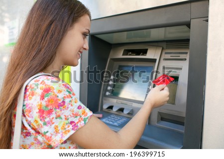 Side portrait view of an attractive young woman using a cash machine to withdraw cash money with her credit card, smiling in a city street. Outdoors finance and lifestyle.