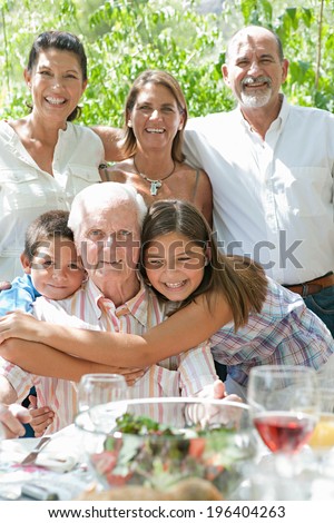 Portrait of a happy family with joyful children hugging their grandfather during a sunny summer day on a holiday home green garden while having lunch outdoors. Family relaxing on vacation, lifestyle.