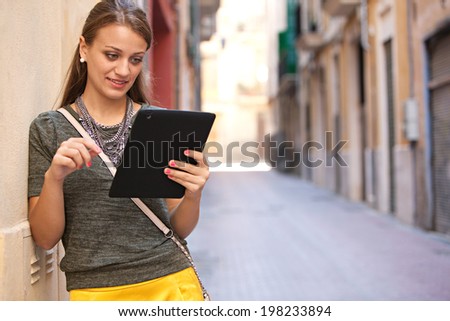 Close up portrait of an attractive young woman leaning on a wall using a digital tablet while visiting a characterful city street and smiling during a sunny day outdoors. Technology and lifestyle.