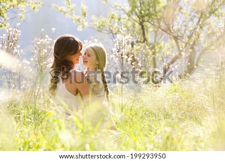 Young mother and child daughter together, hugging and joyfully laughing while relaxing in a golden field of sunshine and spring flowers while on a summer holiday. Family outdoors lifestyle.