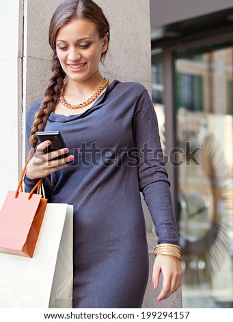 Portrait of a smart young woman shopping by an elegant store entrance in the city and using her smartphone cell during a visit to a luxurious shopping mall. Consumerism and technology, outdoors.