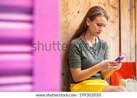 Close up portrait of an attractive young woman sitting on an old aged wooden door with her shopping bags, being thoughtful and using a smartphone to go on line, outdoors. Technology and lifestyle.