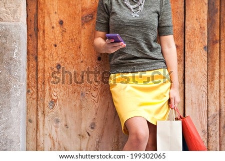 Middle section view of a young woman legs carrying shopping bags and using a smartphone to connect on line and network during a summer day. Tourism, technology and consumerism outdoors.