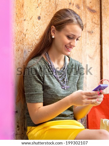 Close up portrait of an attractive young woman sitting by an old textured wooden door with shopping bags, joyfully smiling using a smartphone to network on vacation. Outdoors technology and lifestyle.