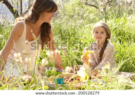 Close up portrait of a young mother and daughter relaxing together having a picnic in a lush green garden eating a healthy fresh banana. Family activities and healthy eating lifestyle, outdoors.