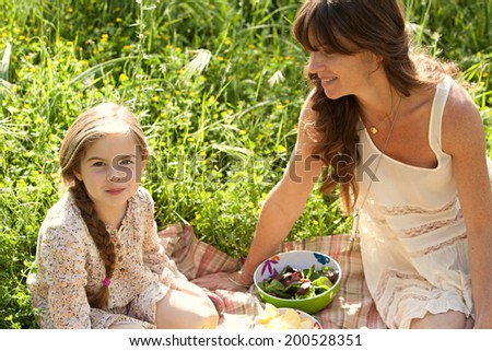 Close up portrait of a young mother and daughter relaxing together having a picnic in a lush green garden with long grass and golden sunshine. Family activities and healthy eating lifestyle, outdoors.