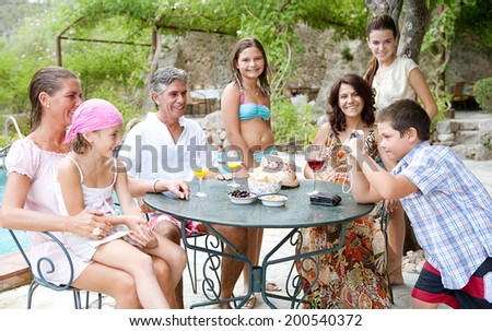 Large family group relaxing together at a table with food and drink in a summer holiday hotel garden with swimming pool, posing for a photograph during a vacation together. Active family lifestyle.