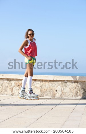 Active african american black teenager girl exercising and roller skating by the beach, smiling at the camera during a summer sunny day on vacation, outdoors. Attractive girl against a blue sky.