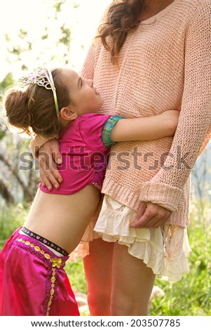 Side view of a pretty young girl wearing a pink fancy dress, hugging her mothers legs while having fun and enjoying a sunny holiday in a green park field, outdoors. Active family lifestyle.