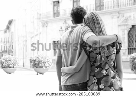 Black and white rear view of a young tourist couple relaxing together and sightseeing in a destination city square while on a summer holiday, outdoors. Travel lifestyle and discovery vacation.