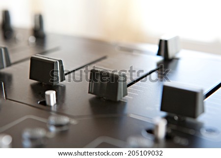 Close up detail view of a DJ mixing deck with buttons and control slides against a bright light, recording studio interior. Music equipment and professional sampling technology.