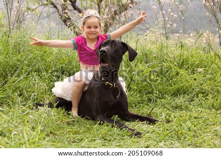 Young child girl dressing up in a fancy dress, sitting on her great dane dogs back holding her arms up like a heroine. Proud dog owner playing with her pet and enjoying a summer holiday together.